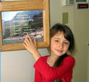 A young girl named Katie, pointing to her name on the Assessments appointment listing at DCC.