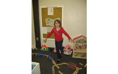 Katie standing in the family room next to a doll house, baby doll stroller, toy barn, and toy train tracks.